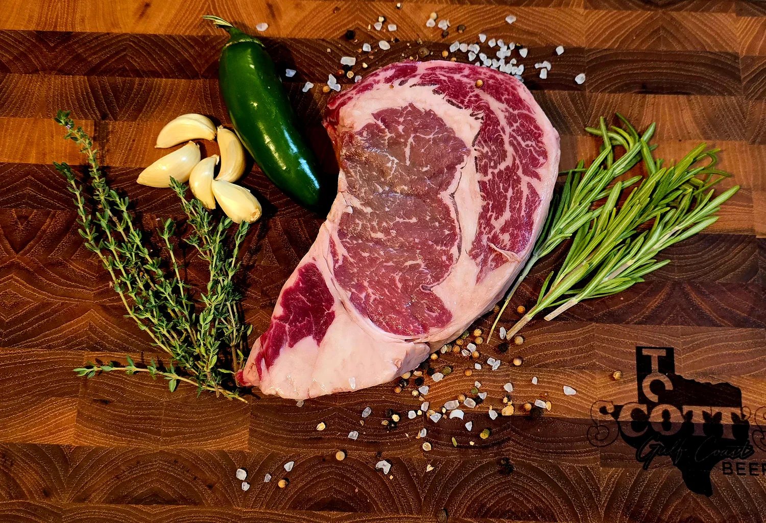 A beautifully marbled ribeye steak surrounded by fresh garlic cloves, a jalapeño pepper, sprigs of thyme and rosemary, and a sprinkle of coarse salt and pepper on a wooden cutting board.