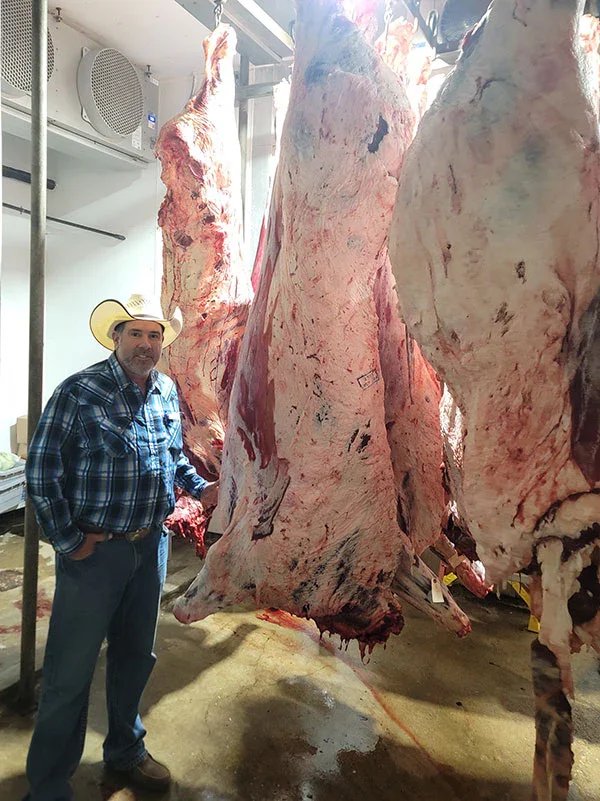 Man wearing a cowboy hat and plaid shirt stands next to hanging beef carcasses in a meat processing facility, showcasing the butchering and meat preparation process.