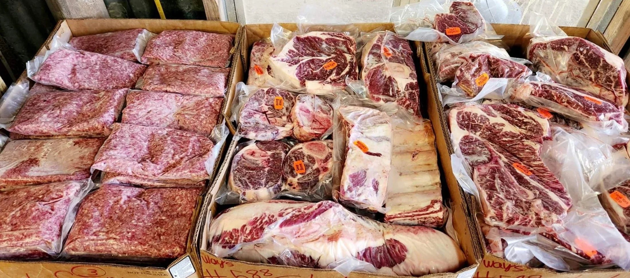 Assorted packaged meats displayed in cardboard boxes, including ground beef, ribeye steaks, and various cuts, with visible price tags and labels for easy identification.