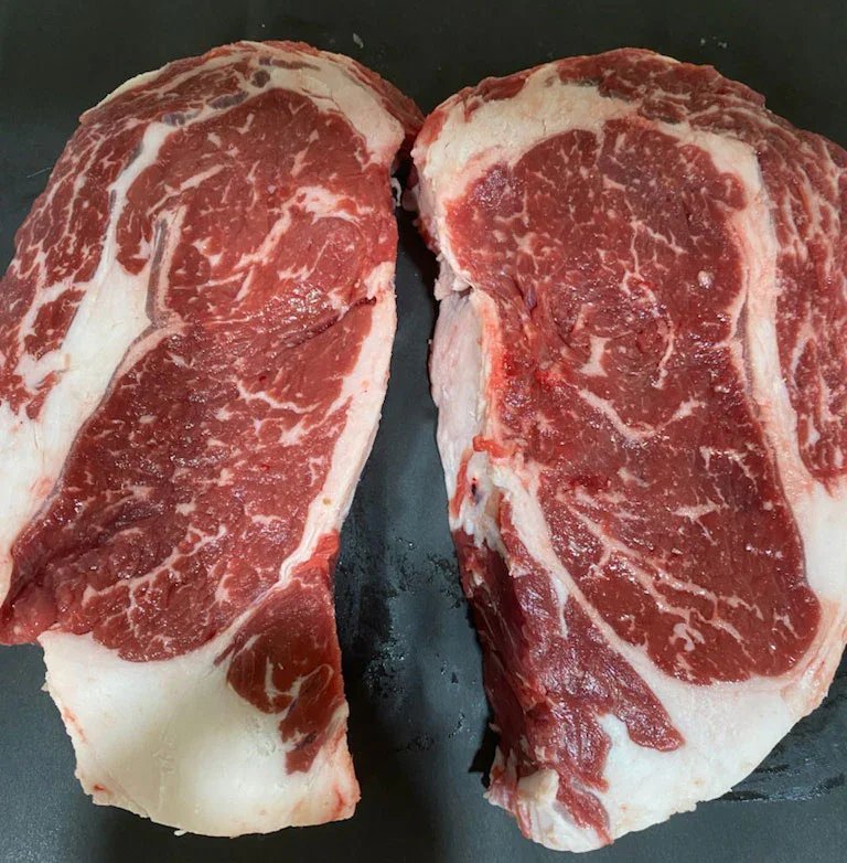 Close-up image of two raw ribeye steak cuts on a black surface, showcasing marbling and rich red color, ideal for grilling or roasting.