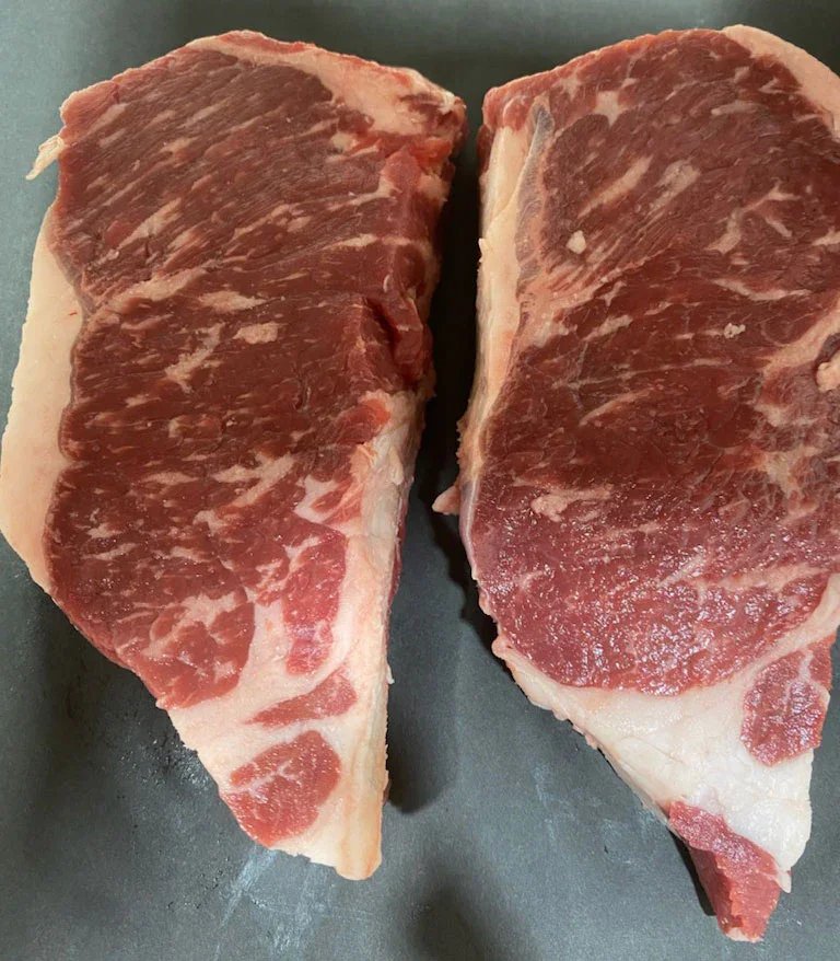 Two raw ribeye steak cuts displayed on a dark surface, showcasing marbling and a thick layer of fat for enhanced flavor and tenderness. Ideal for grilling or pan-searing.