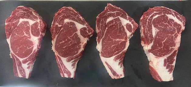 Four marbled ribeye steaks displayed on a black cutting board, showcasing rich red meat and white fat marbling, ideal for grilling or roasting.