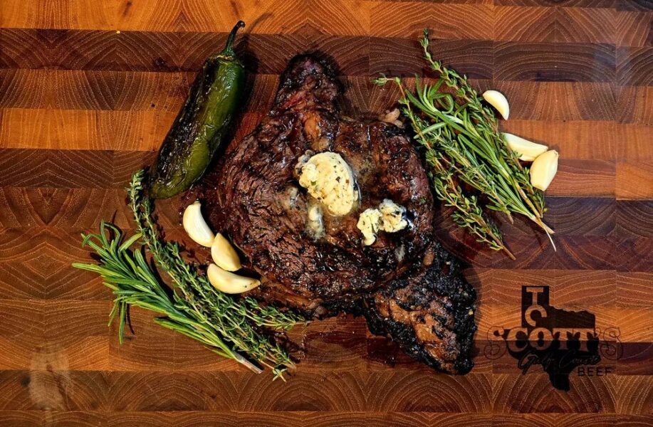 Juicy grilled ribeye steak topped with herb butter, surrounded by fresh garlic cloves, rosemary, thyme, and a green jalapeño pepper, presented on a wooden cutting board. Ideal for steak lovers and culinary enthusiasts.