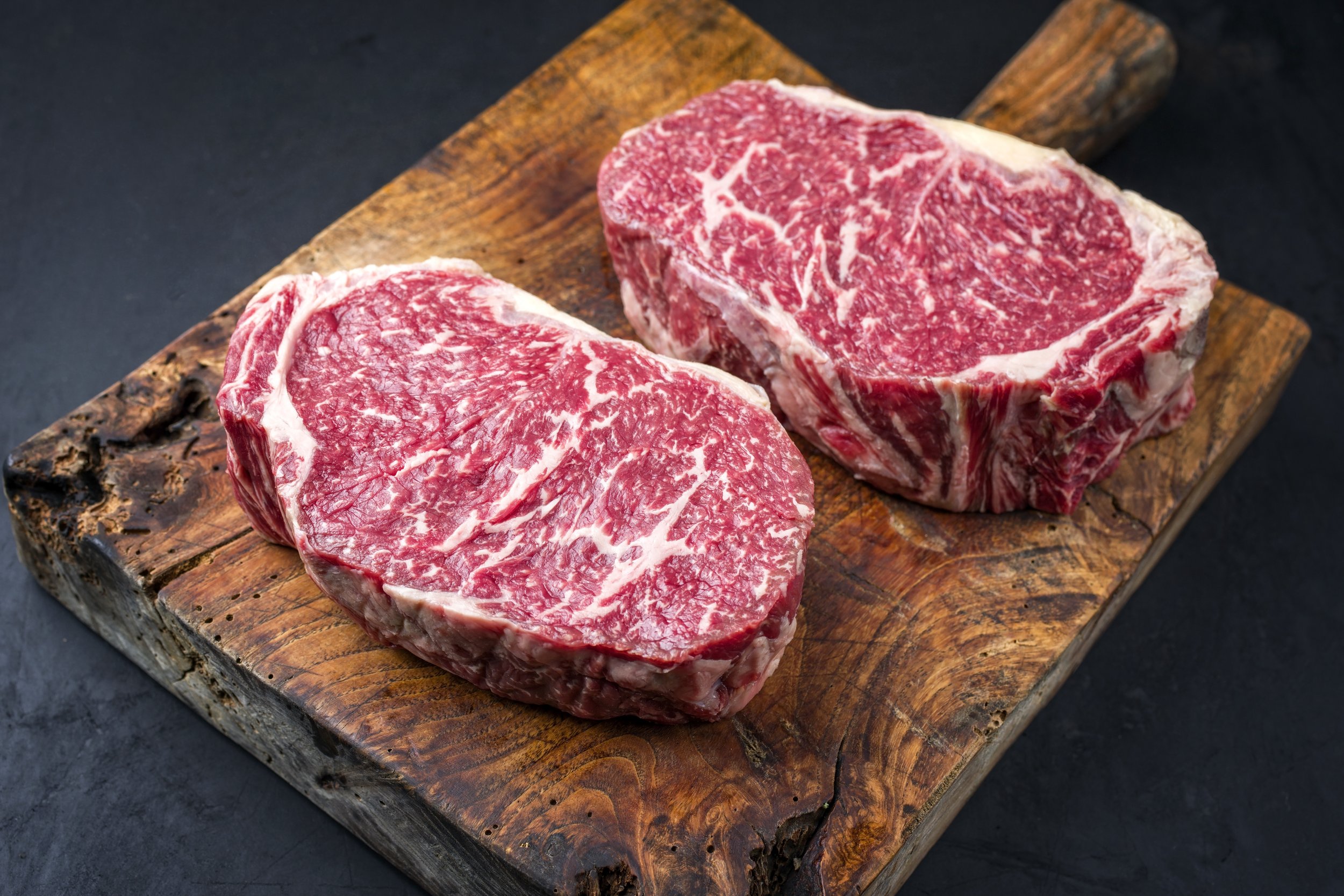 Two raw ribeye steaks with rich marbling, displayed on a rustic wooden cutting board, highlighting their premium quality and texture.
