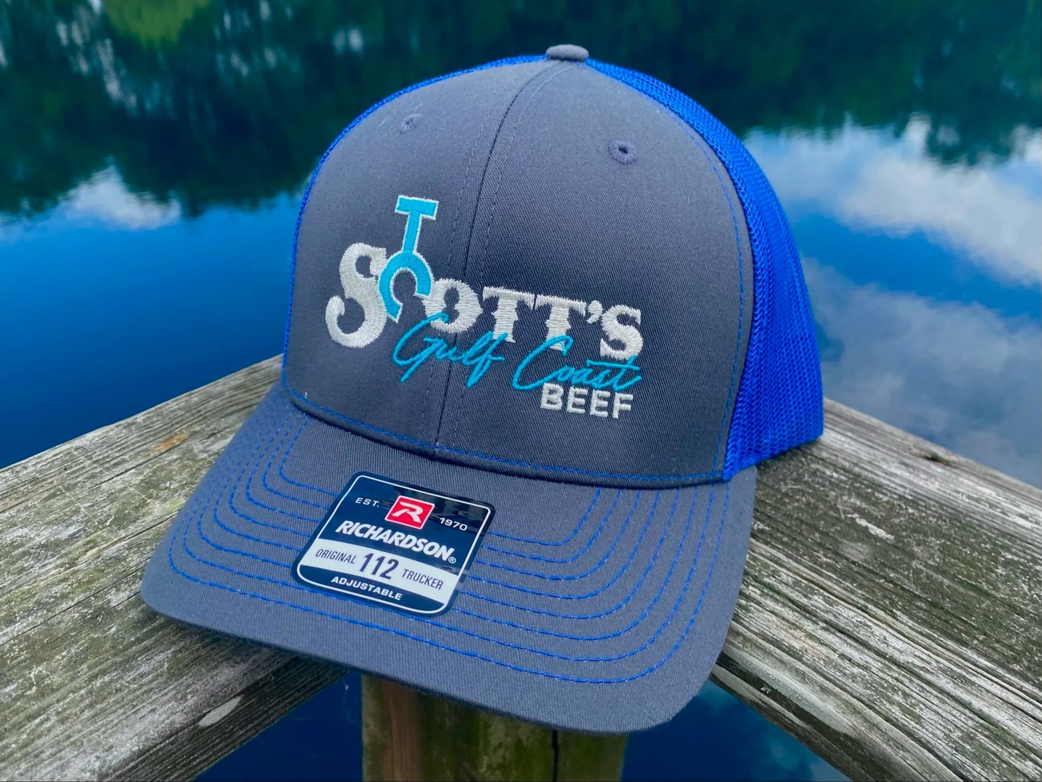 Gray and blue Richardson 112 trucker hat featuring "Scott's Gulf Coast Beef" logo, resting on a wooden surface with a serene lake background.