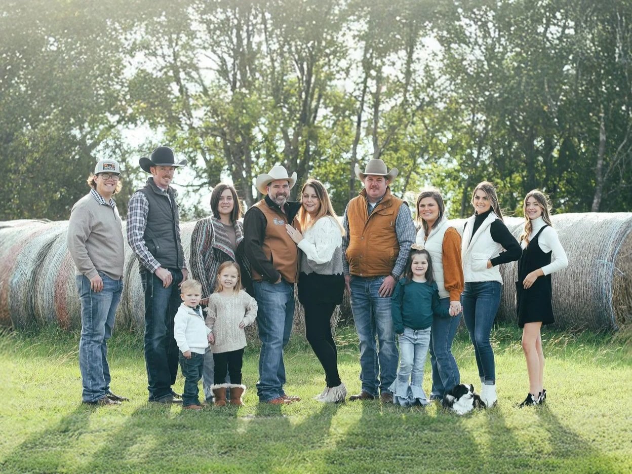 A large group of people, including men, women, and children, pose together outdoors near hay bales in a rural setting. The adults are dressed in casual and western-style clothing, while children playfully stand in front. The background features trees and greenery, creating a warm, family-oriented atmosphere.