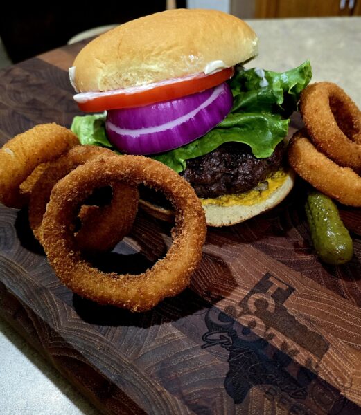 Juicy hamburger with lettuce, tomato, red onion, and mayonnaise on a toasted bun, accompanied by crispy onion rings and pickles, served on a wooden cutting board.